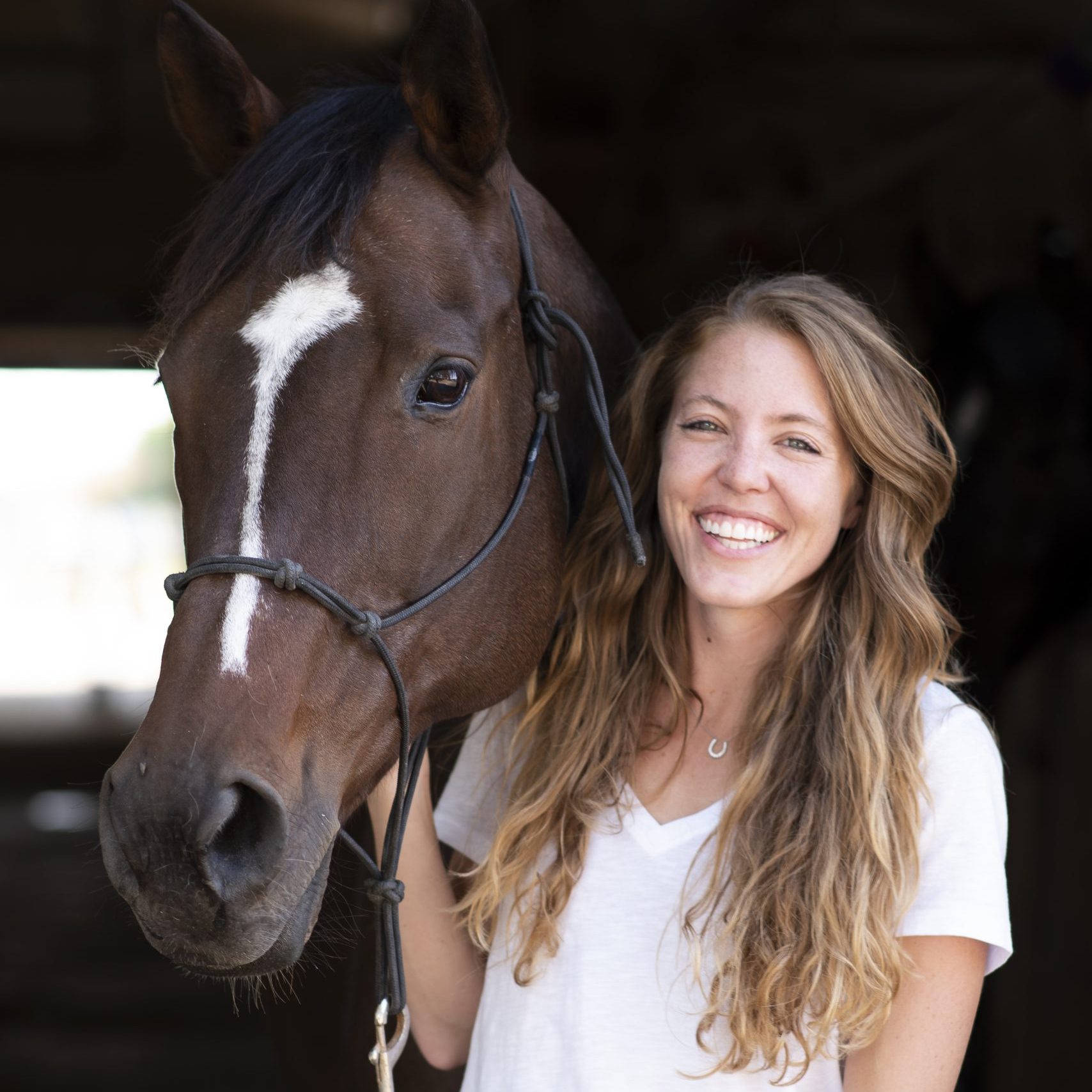 Smiling girl stands next to her bay horse with a white stripe on her face in a dark barn isle.