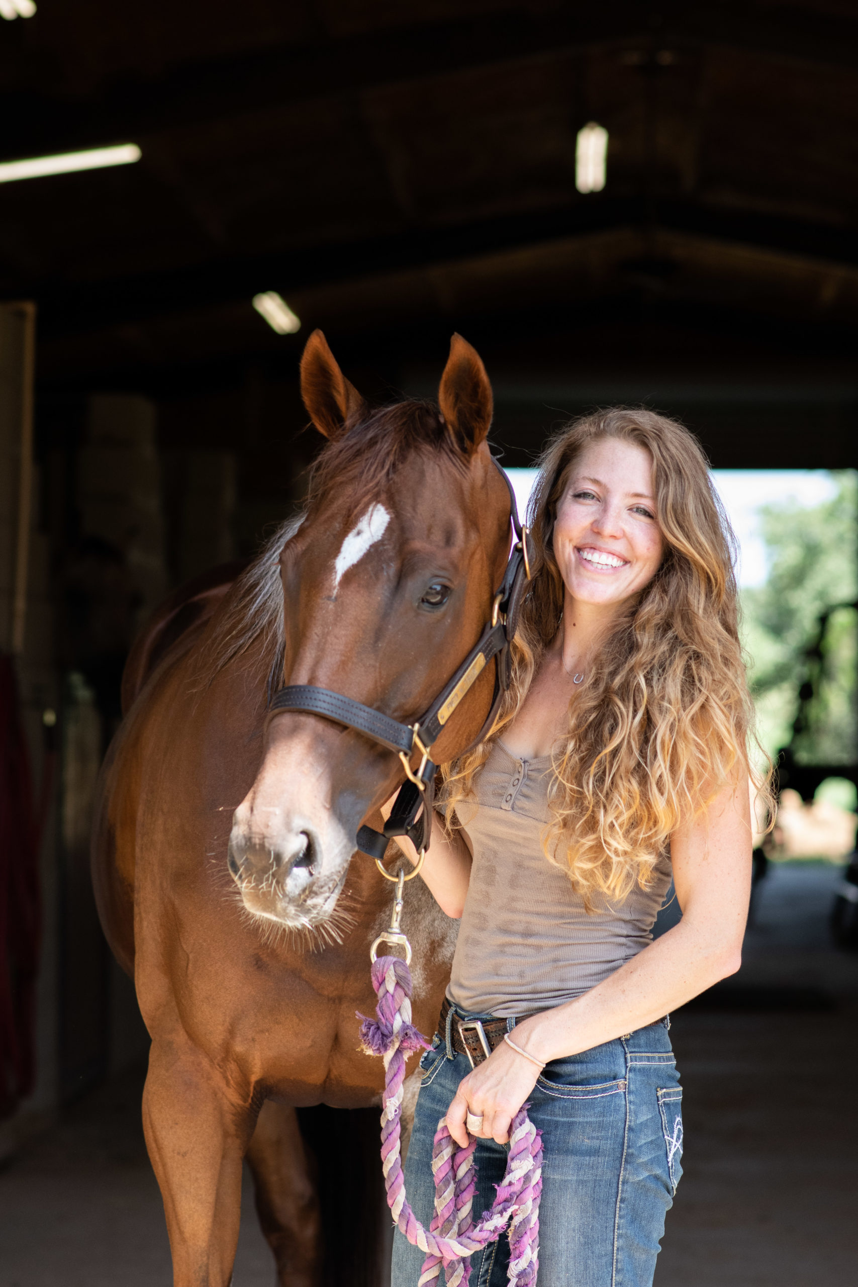 A smiling girl stands next to her sorrel horse in a barn isle