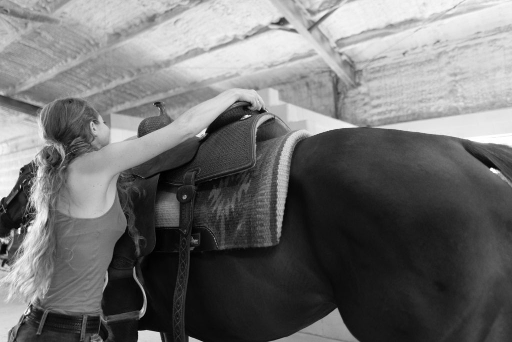 A girl swings her Western saddle up over her horse's back