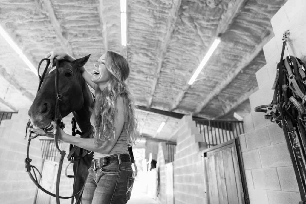 A smiling girl brides her horse in a barn isle