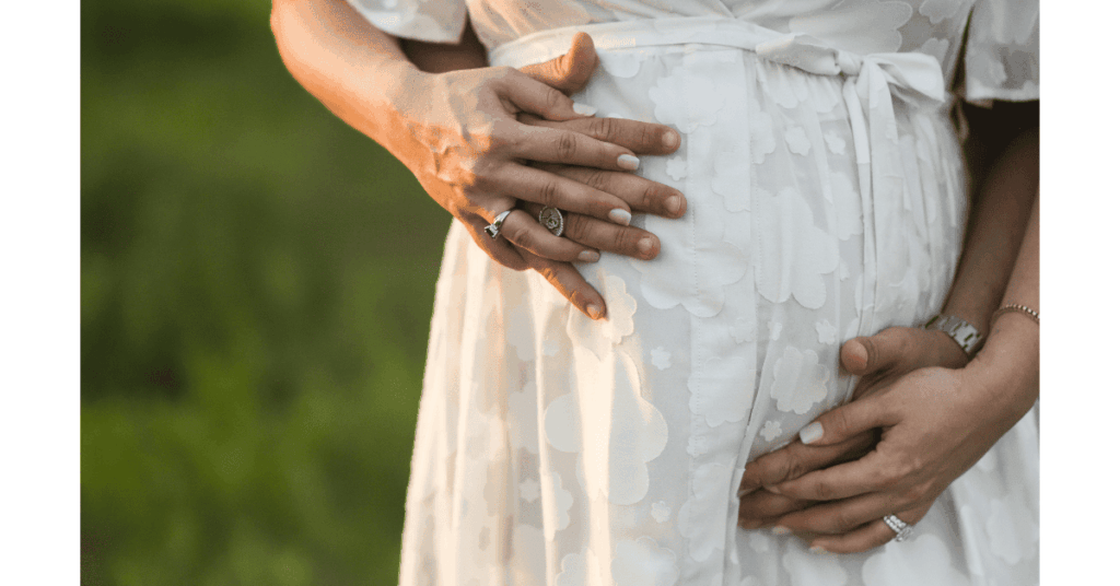 Relaxed, documentary-style maternity photo of a mom-to-be in a dress with her husband's tender arms around her cradling their daughter
